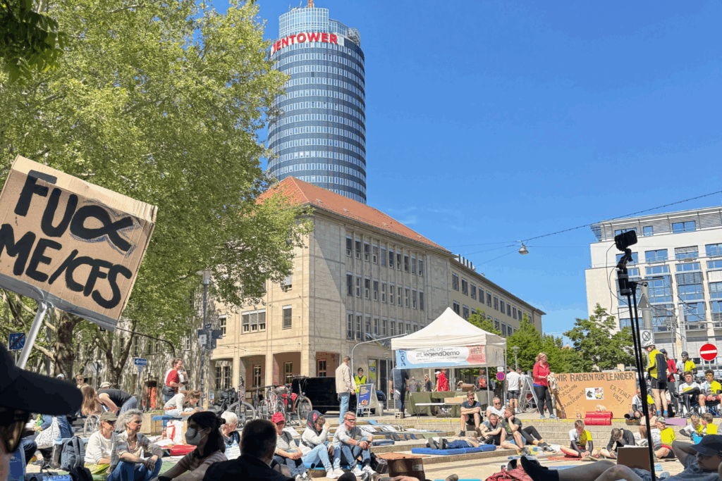 Ein großes Schild im Vordergrund mit der Aufschrift „FUCK ME/CFS“ sticht hervor. Im Hintergrund ist der JenTower und ein großer weißer Pavillon mit der Aufschrift „#Liegenbleiben“ zu sehen. Viele Menschen liegen oder sitzen auf dem Platz, einige auf Isomatten oder Decken. Es handelt sich um die Liegenddemo 2025 in Jena zur Sensibilisierung für ME/CFS, bei der das Liegenbleiben symbolisch für die Krankheit steht. Die Atmosphäre wirkt friedlich, bei sonnigem Wetter.