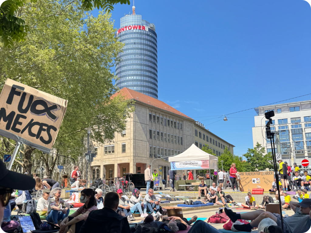 Ein großes Schild im Vordergrund mit der Aufschrift „FUCK ME/CFS“ sticht hervor. Im Hintergrund ist der JenTower und ein großer weißer Pavillon mit der Aufschrift „#Liegenbleiben“ zu sehen. Viele Menschen liegen oder sitzen auf dem Platz, einige auf Isomatten oder Decken. Es handelt sich um die Liegenddemo 2025 in Jena zur Sensibilisierung für ME/CFS, bei der das Liegenbleiben symbolisch für die Krankheit steht. Die Atmosphäre wirkt friedlich, bei sonnigem Wetter.