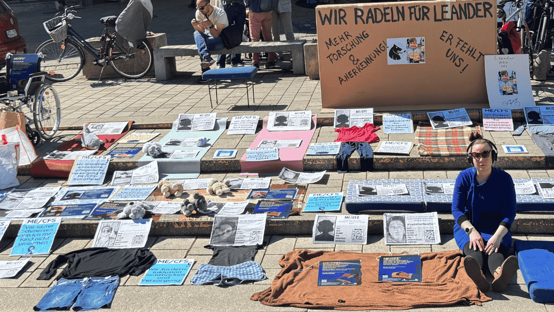 Auf den Stufen des Holzmarktes in Jena repräsentieren ausgelegte Kleidungsstücke, Decken, Stofftiere und Steckbriefe die vielen an ME/CFS Erkrankten, die nicht persönlich auf der Liegenddemo anwesend sein können. Im rechten Bereich des Bildes sitzt eine junge Frau mit Kopfhörern allein auf einer der Stufen. Sie trägt ein blaues Langarmshirt und blickt direkt in die Kamera. Ihr Gesichtsausdruck wirkt ernst und entschlossen.