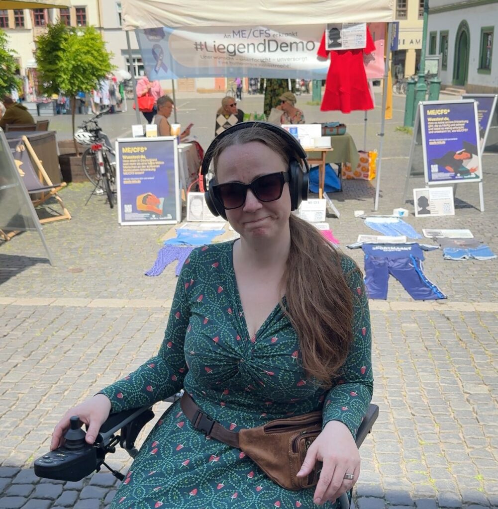 Eine junge Frau mit langen braunen Haaren, Sonnenbrille und Kopfhörern sitzt in einem elektrischen Rollstuhl und blickt in die Kamera. Sie befindet sich im Freien auf einem gepflasterten Platz. Im Hintergrund sind Stände und Plakate der Initiative #LiegendDemo zu sehen, die auf die chronische neuroimmunologische Erkrankung ME/CFS (Myalgische Enzephalomyelitis/Chronisches Fatigue-Syndrom) aufmerksam machen soll. Auf dem Boden vor den Ständen liegen Kleidungsstücke, die symbolisch die Anwesenheit von Betroffenen repräsentieren, die zu krank zum Demonstrieren sind.