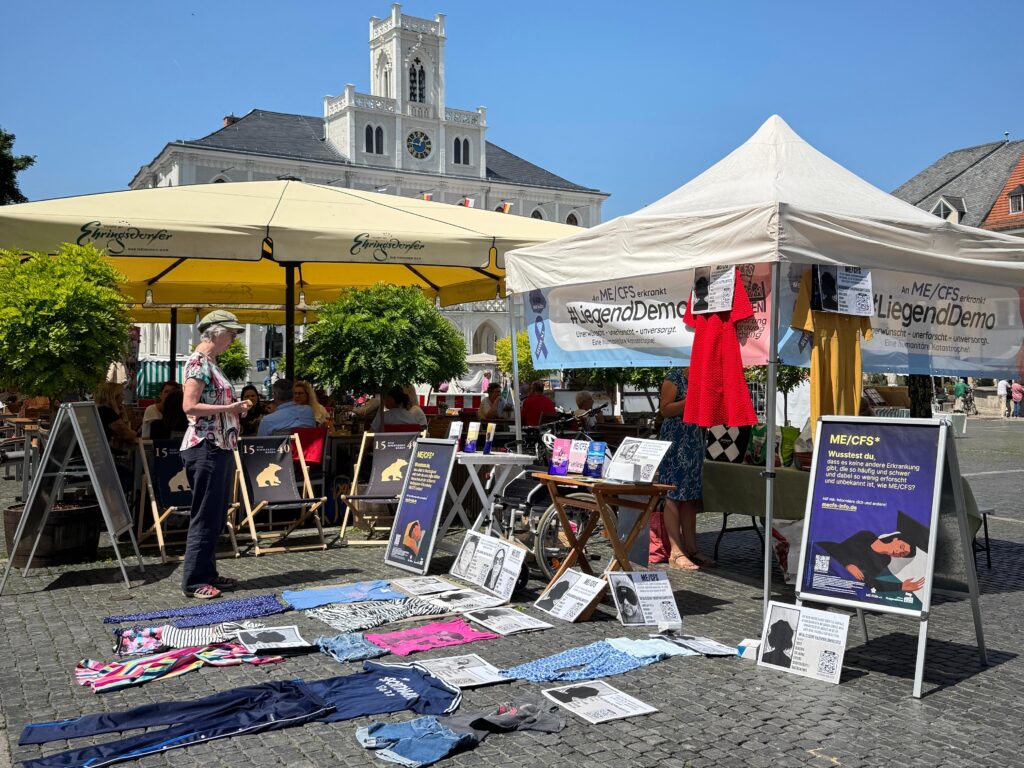 Das Bild zeigt einen Informations- und Proteststand der Initiative #LiegendDemo auf einem gepflasterten Marktplatz, im Hintergrund ist das markante weiße Rathaus von Weimar zu sehen. Im Vordergrund liegen am Boden Kleidungsstücke, die symbolisch für die „liegende“ Demonstration stehen. Eine Passantin ist stehengeblieben und betrachtet die ausgelegten Steckbriefe Betroffener. Am und neben dem Infostand hängen weitere Infoblätter.