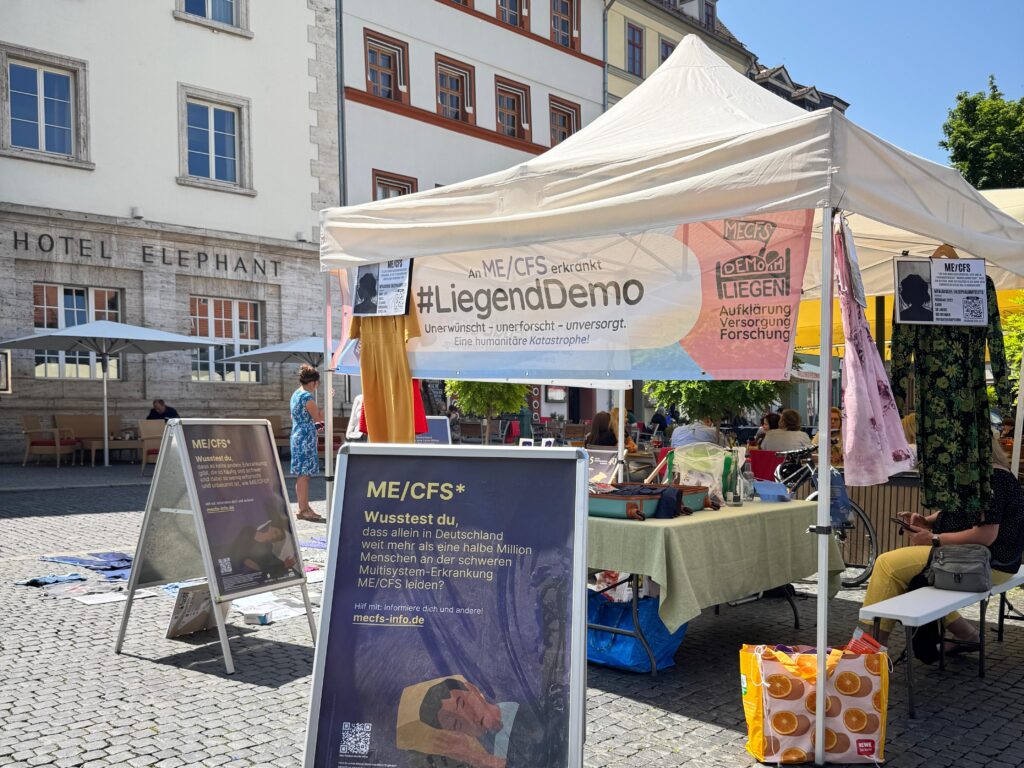 Ein heller Informationsstand der Initiative #LiegendDemo steht auf dem gepflasterten Marktplatz in Weimar, direkt vor der markanten Fassade des historischen Hotel Elephant. An der Front ist ein großes Banner mit der Aufschrift „#LiegendDemo – An ME/CFS erkrankt. Unerwünscht – unerforscht – unversorgt. Eine humanitäre Katastrophe!“ befestigt. Im Vordergrund steht ein großer Aufsteller mit einem Foto einer liegenden Person und dem Text: „Wusstest du, dass allein in Deutschland weit mehr als eine halbe Million Menschen an der schweren Multisystem-Erkrankung ME/CFS leiden?“. Ein QR-Code verweist auf weitere Informationen. Am Infostand hängen weitere Infoblätter. Am Boden liegen Kleidungsstücke, die symbolisch für die „liegende“ Demonstration stehen könnten.