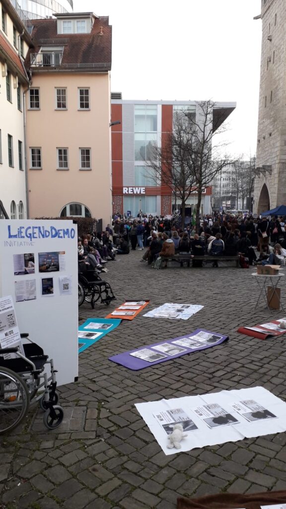 Das Foto zeigt eine öffentliche Versammlung auf einem gepflasterten Stadtplatz. Im Vordergrund steht eine große weiße Informationstafel mit der blauen Aufschrift „LIEGENDDEMO INITIATIVE“. Daneben ist ein leerer Rollstuhl platziert. Auf dem Boden sind mehrere bunte Isomatten ausgelegt. Auf jeder Matte kleben Plakate mit Schwarz-Weiß-Porträts und kurzen Texten, die Einzelschicksale von an ME/CFS erkrankten Menschen beschreiben. Ein kleiner weißer Plüschhase liegt symbolisch auf einem der vorderen Plakate.