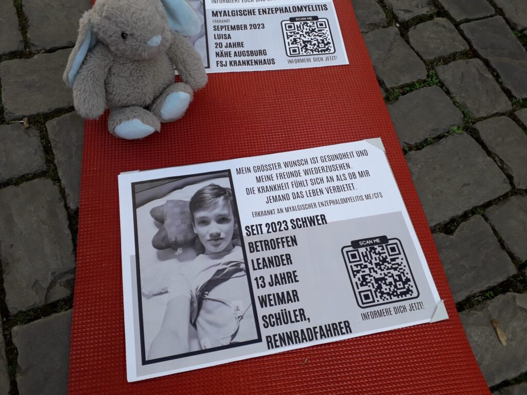 Zwei Informationsplakate, die auf einer roten Isomatte auf einem Kopfsteinpflasterboden liegen. Oben links sitzt ein kleiner, grauer Plüschhase. Die Plakate machen auf die Krankheit Myalgische Enzephalomyelitis (ME/CFS) aufmerksam und zeigen persönliche Schicksale. Das untere Plakat im Fokus zeigt ein Schwarz-Weiß-Foto eines 13-jährigen Jungen namens Leander aus Weimar, der im Bett liegt. Daneben steht: „Mein größter Wunsch ist Gesundheit und meine Freunde wiederzusehen. Die Krankheit fühlt sich an als ob mir jemand das Leben verbietet.“ Er wird als Schüler und Rennradfahrer beschrieben, der seit 2023 schwer betroffen ist.